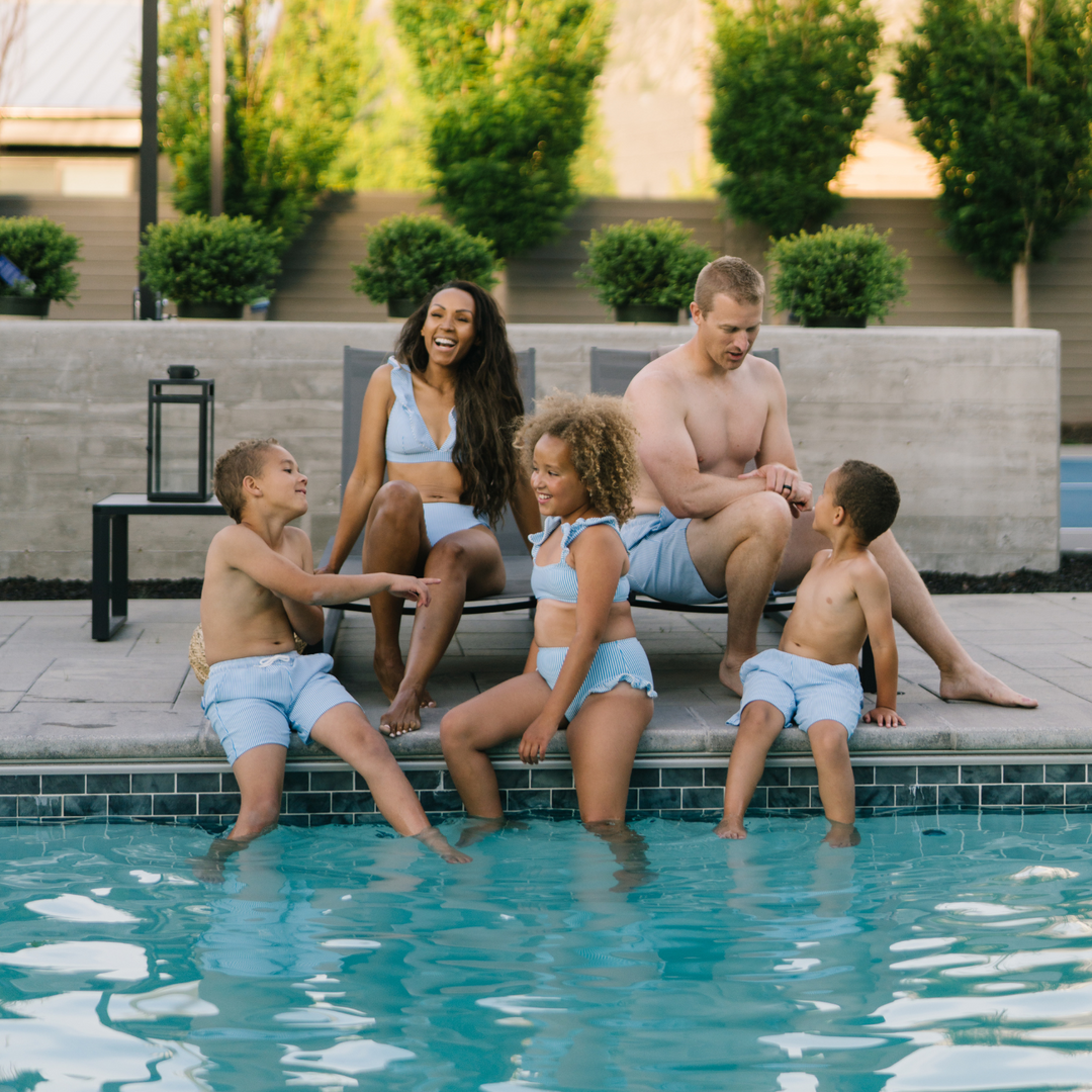 Family sitting poolside wearing matching seersucker swimsuits.  The mom and daughter have matching bikinis and the Dad and son's are wearing matching swim shorts.